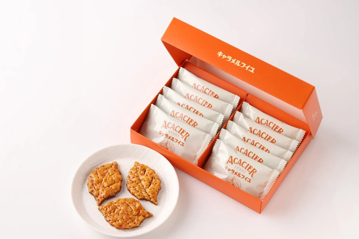 オレンジ色の箱に入ったお菓子。箱には「ACACIER」の文字と「キャラメルフイユ」の文字が書かれている。箱の横には、同じお菓子が白い皿に3つ置かれている。