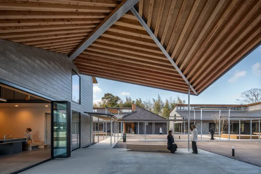 木材とコンクリートで作られた建物の外観。屋根は木材でできており、人々が座って会話をしている様子が見える。周囲には他の建物や木々が見える。