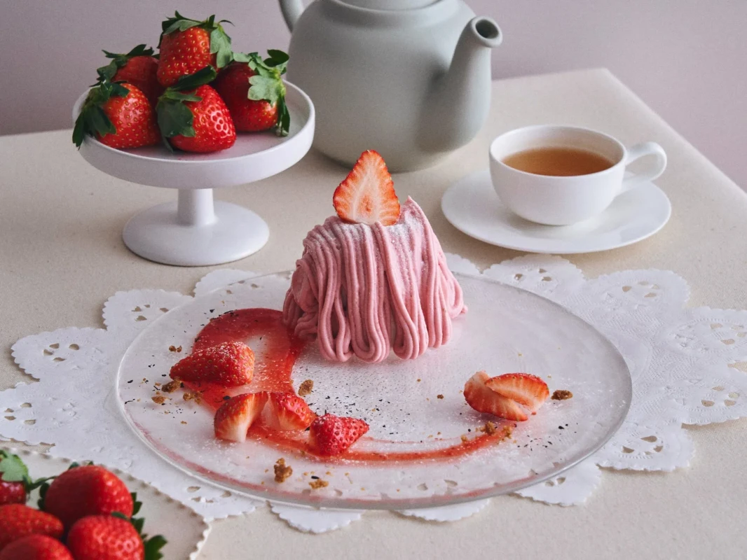 いちごのモンブランケーキと紅茶のティーセットの写真。モンブランケーキはピンク色のクリームで覆われ、上にいちごのスライスが乗っている。周りにはカットされたいちご、いちごソース、ティーカップ、ティーポットが配置されている。