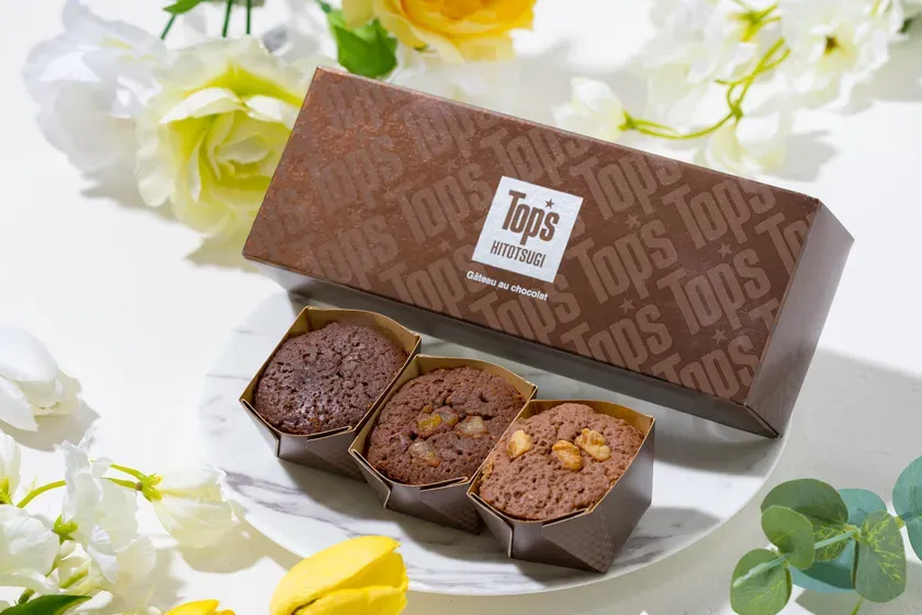 茶色の箱に入ったチョコレートケーキと、白い皿の上に載せられた3つのチョコレートケーキ、そして周りには花が飾られた写真です。