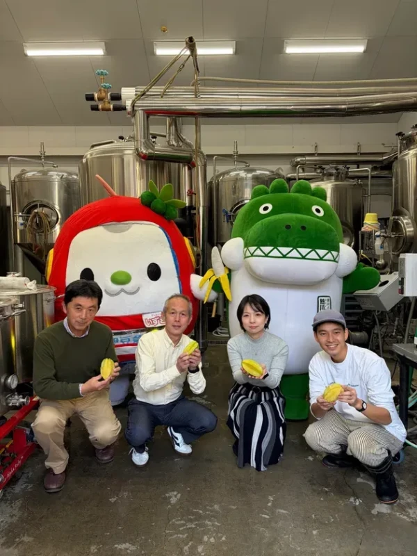 4人の人々が、2つのマスコットと一緒に、スターフルーツのような果物を持っています。人々は醸造所のような場所にいます。