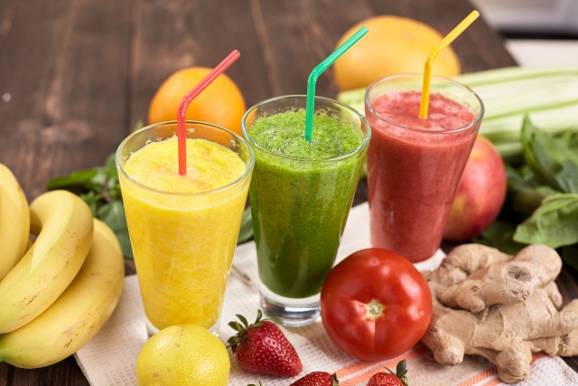 野菜スムージー3種 黄色と緑と赤 トマトやイチゴ、レモン、オレンジが背景に置かれている