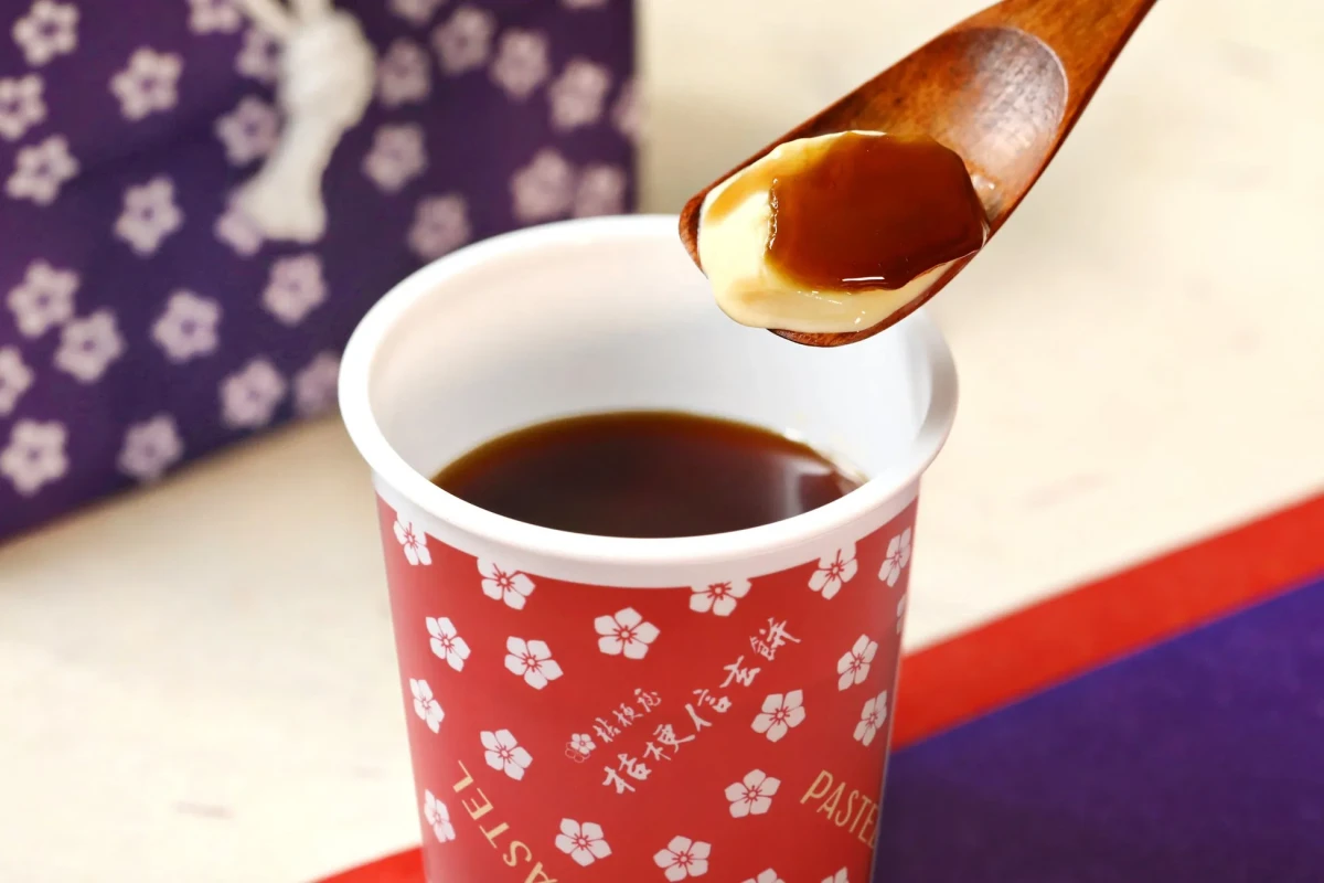 赤いカップに入ったプリンを木製スプーンですくっている様子を捉えた写真です。カップには花柄と「桔梗信玄餅 PASTEL」の文字が見えます。背景には紫色の布がぼかして写っています。