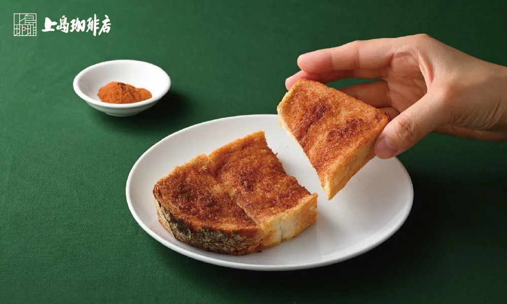 トーストが白い皿に並べられており、手で一つ持ち上げられている写真です。トーストには茶色い砂糖のようなものがまぶされており、白い小皿にはスパイスのようなものが盛られています。