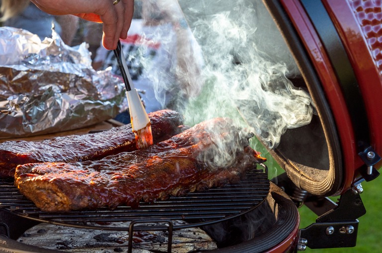 赤いセラミック製バーベキューグリルから煙が立ちのぼり、グリルの上には2ラックのポークリブ。男性がリブにバーベキューソースを塗っている。