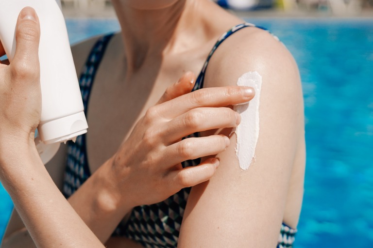 若い女性が、プールの青い水を背景に、白いチューブに入った日焼け止め（SPF入り）を肩に塗っている。スパ、肌の保護、保湿というコンセプトを表現している
